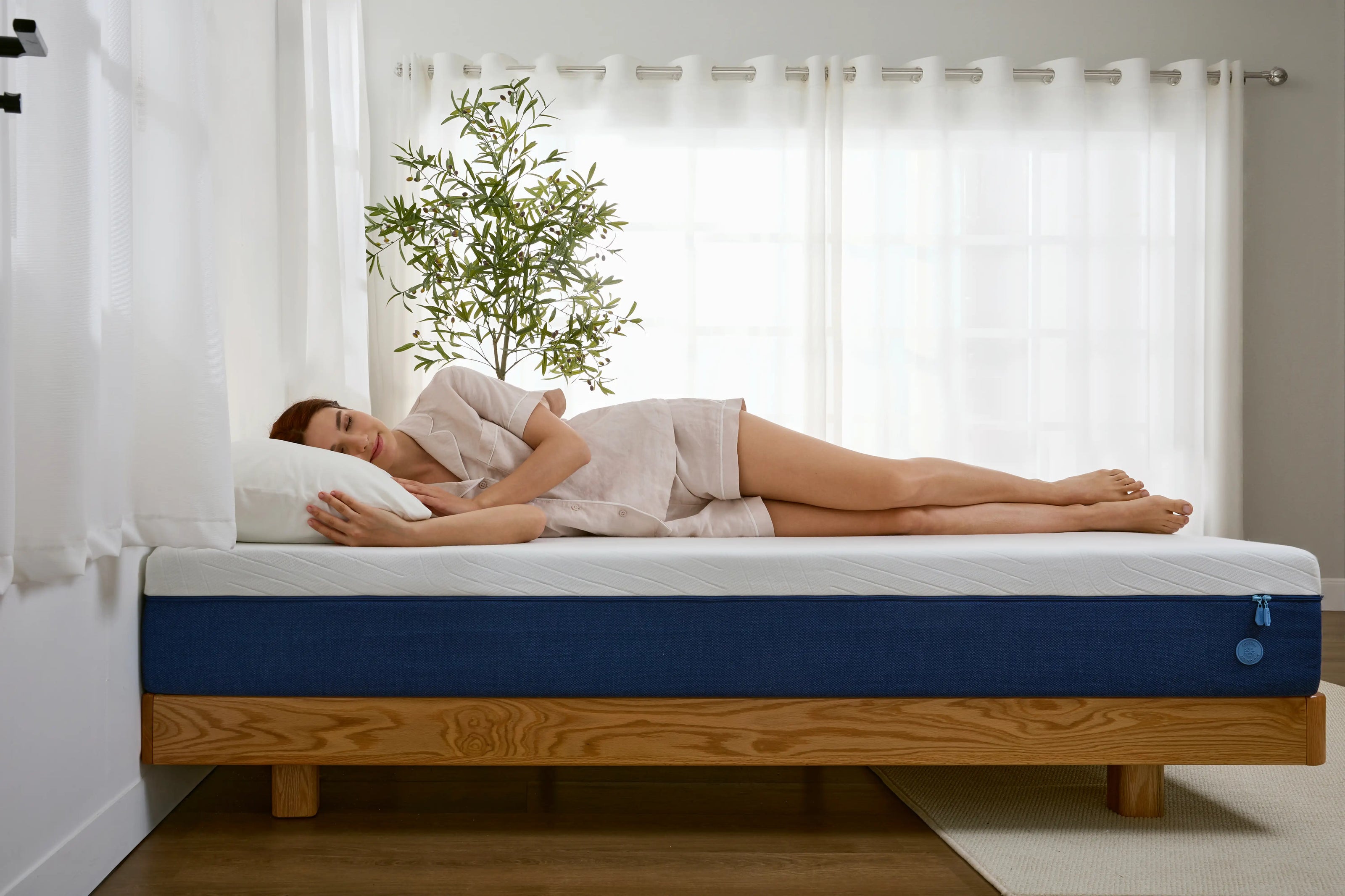 Eine Frau schläft auf der Seite auf einer blauen Matratze mit weißer Oberseite, die auf einem Holzgestell platziert ist. Ihr Körper ist entspannt, und die Matratze zeigt keine starken Einsinkstellen, was sie ideal für Seitenschläfer macht. Im Hintergrund steht eine grüne Zimmerpflanze vor einem hellen Fenster.