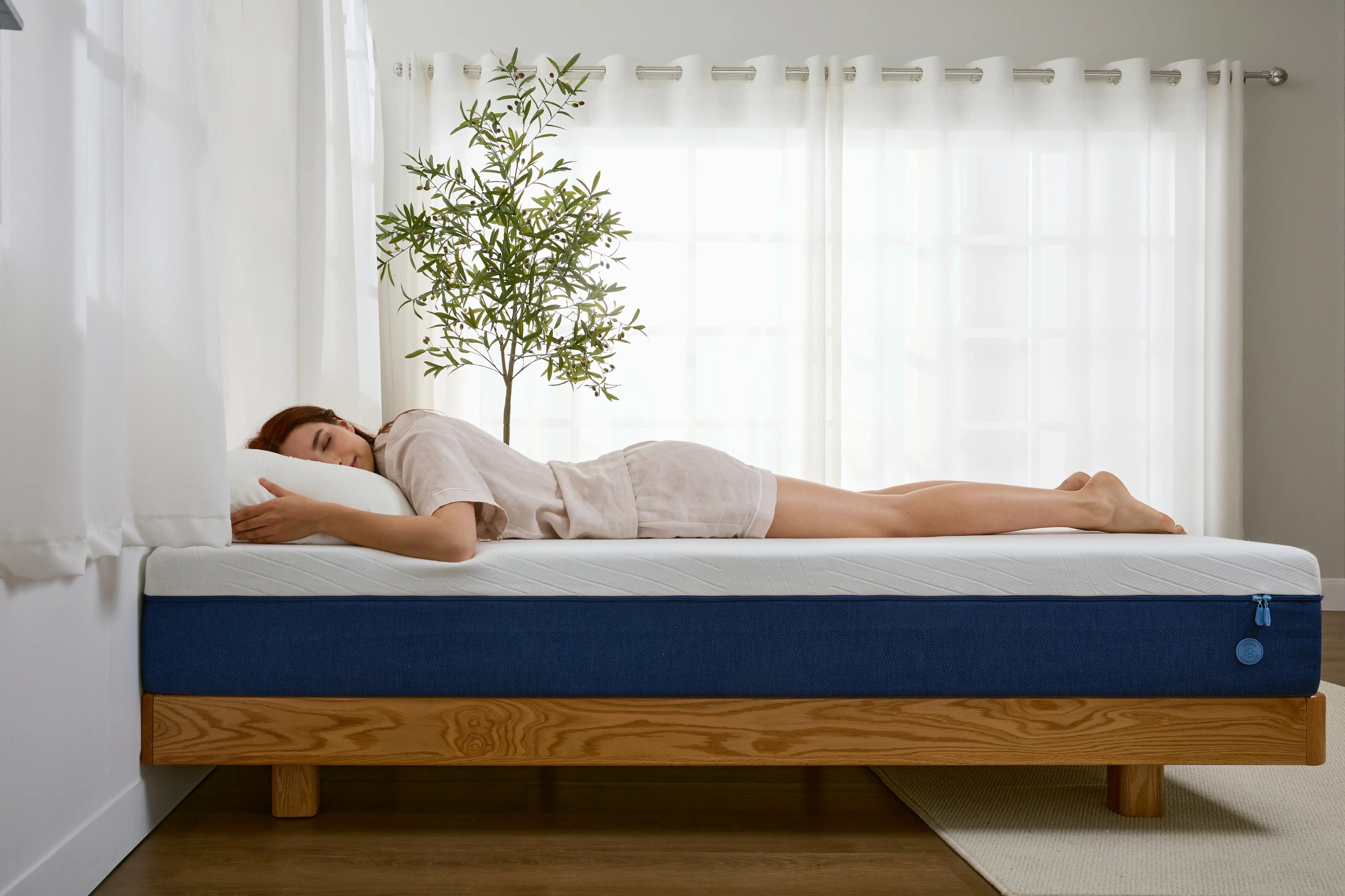 Eine Frau schläft auf dem Bauch auf einer blauen Matratze mit weißer Oberseite, die auf einem minimalistischen Holzgestell platziert ist. Im Hintergrund steht eine grüne Zimmerpflanze vor einem hellen Fenster mit Vorhängen. Diese Ansicht zeigt die Stabilität und Ebenheit der Matratze, ideal für Bauchschläfer.